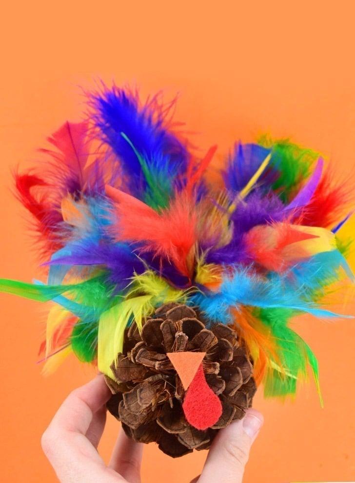 Fall craft for kids, a hand holding a pinecone turkey with rainbow craft feathers, an orange felt beak and a red wattle