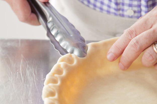 Hands crimping the raised edge of an unbaked pie crust with metal kitchen tongs on a wooden work surface