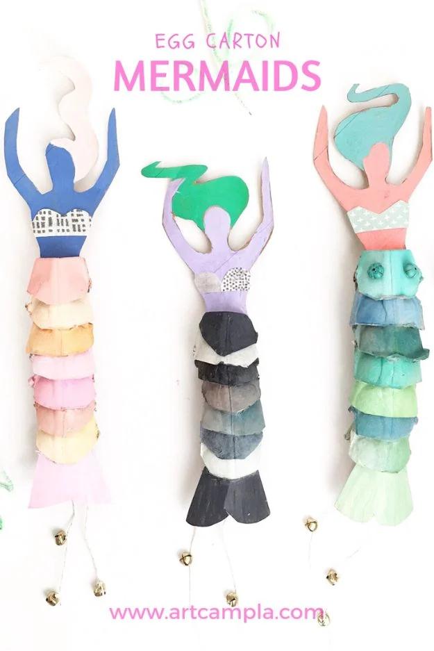 Three egg carton mermaid dolls with stacked scale tails in pink, gray and green, arms raised, hanging against a white wall
