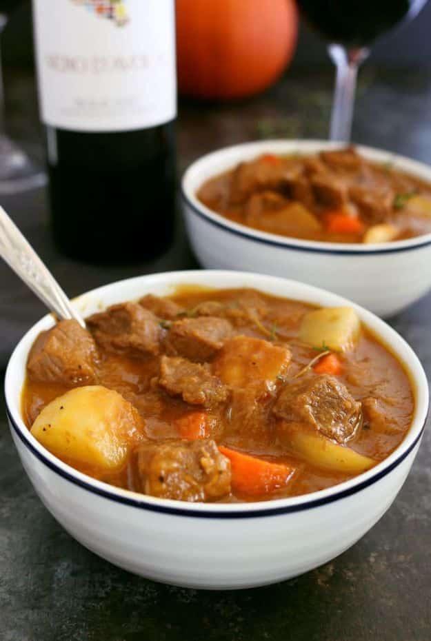 Two bowls of pumpkin beef stew with potatoes and carrots, spoon in one, wine bottle and pumpkin behind