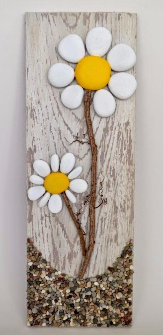 Two daisies made from white pebbles with yellow centers and twig stems on a whitewashed pallet board