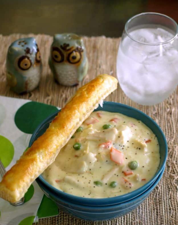 Chicken pot pie soup in a dark blue bowl with a golden puff pastry breadstick resting across the wide rim