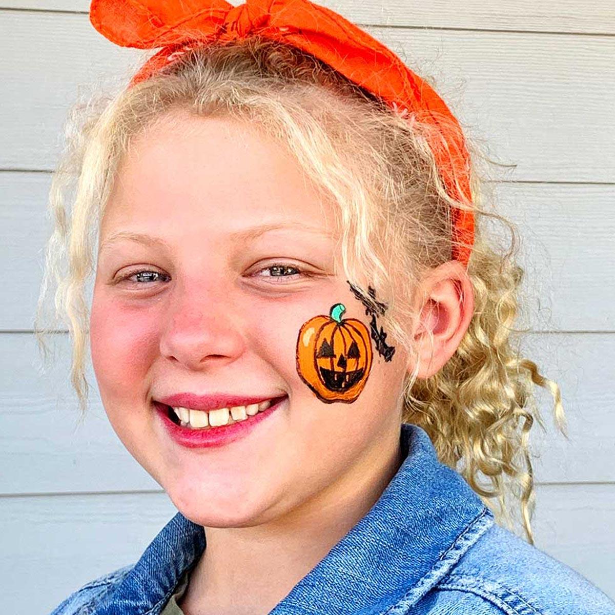 Easy Halloween face paint with a small orange pumpkin and a black bat painted on a smiling girl's cheek