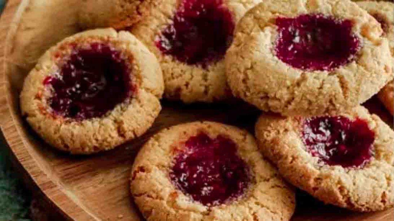 Vegan almond flour thumbprint cookies filled with dark red fruit jam, piled together on a round wooden board