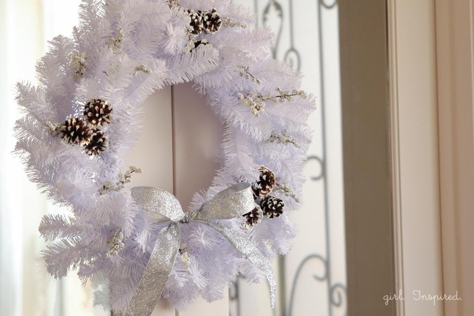 A white wreath dusted with snow, white berry sprigs, snow tipped pinecones and a silver glitter ribbon bow on a glass door