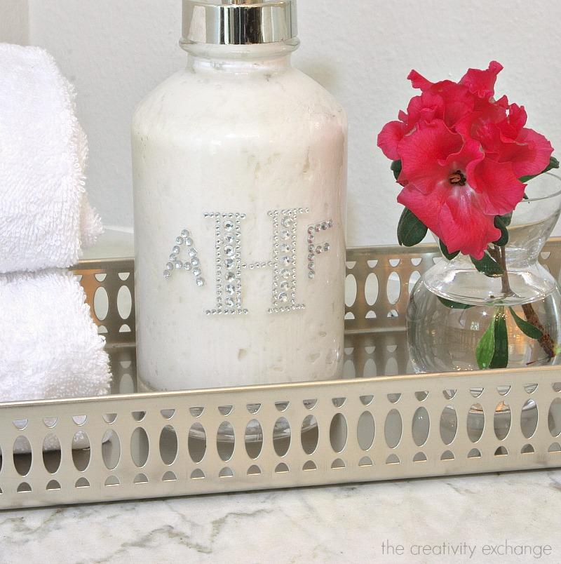 White pump bottle of homemade lotion with a rhinestone AHF monogram on a silver tray beside folded towels and a pink flower
