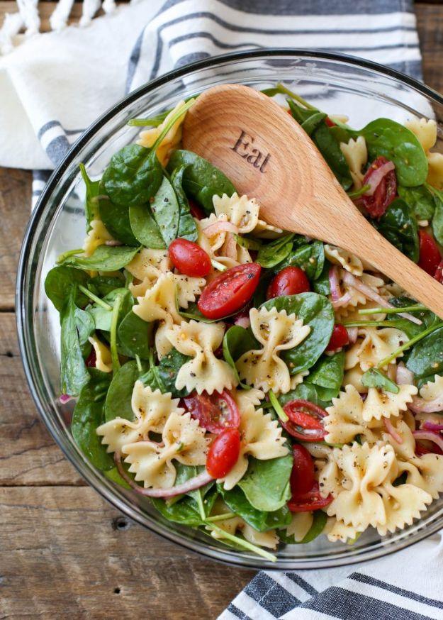 Spinach pasta salad in a glass bowl with bowtie pasta, baby spinach, halved grape tomatoes and a wooden spoon