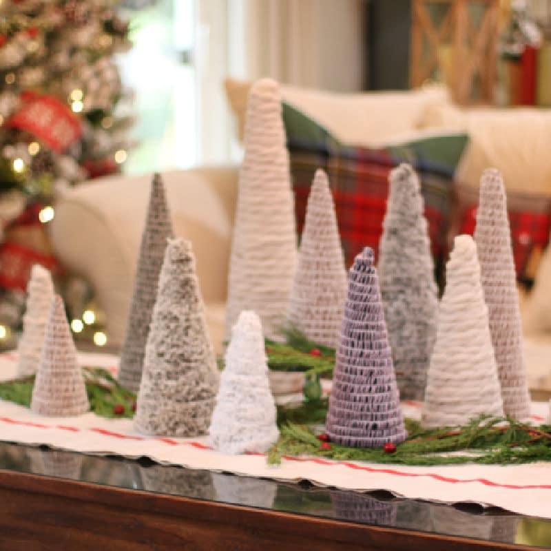 A tabletop forest of cone trees wrapped in cream, gray and violet chunky yarns on a striped runner with fresh greenery