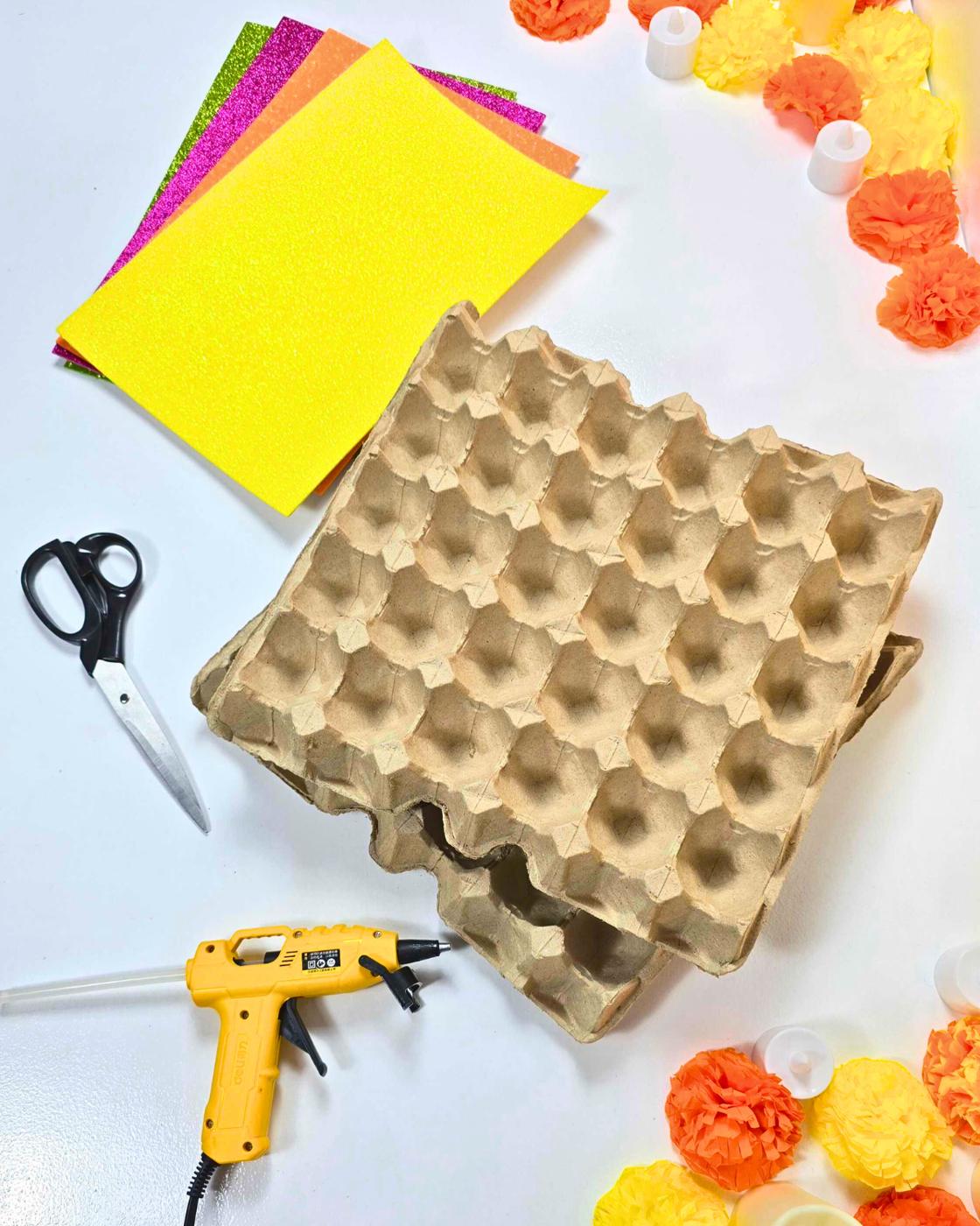 Egg carton crafts supplies - a beige cardboard egg tray with stacked pink, purple, green and yellow glitter foam sheets, scissors, a hot glue gun and paper marigolds