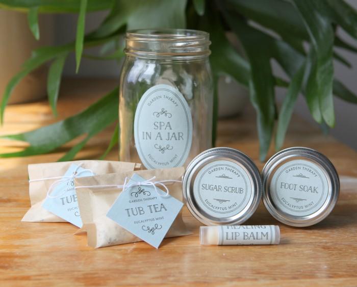 A quart mason jar labeled Spa In A Jar beside tins of sugar scrub and foot soak, kraft packets of tub tea and a lip balm tube