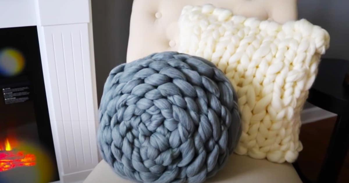 Chunky knit pouf pillows in grey and cream giant yarn, a round one and a square one on a chair by a fireplace