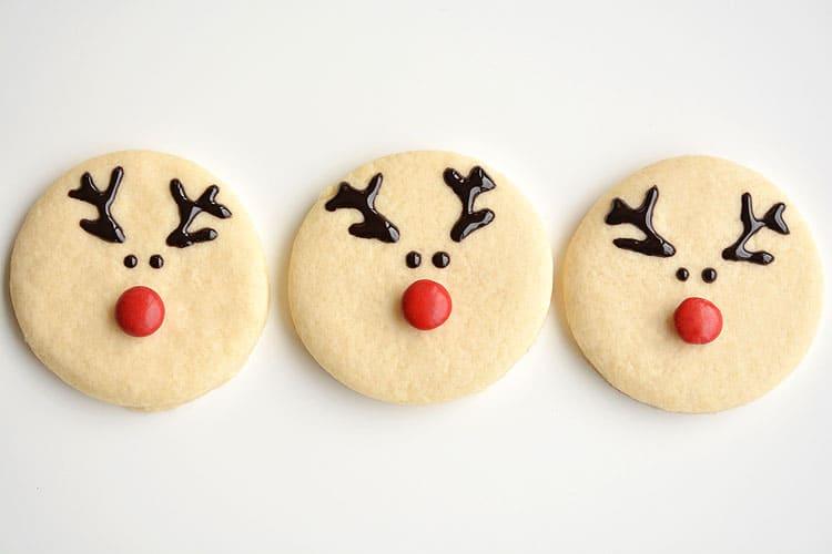 Three round sugar cookies decorated as reindeer with piped chocolate antlers, dot eyes and red candy noses on white