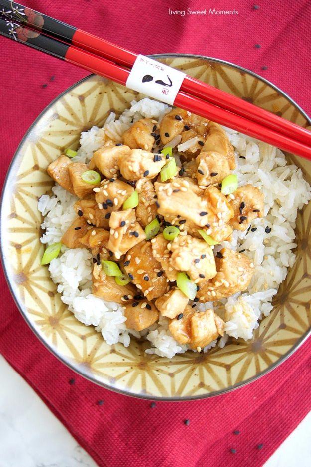 Patterned bowl of sesame chicken over white rice with green onions and sesame seeds beside red chopsticks on red cloth