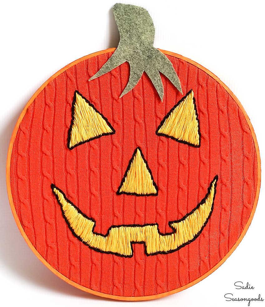 Orange cable knit sweater in an embroidery hoop with a yellow jack-o-lantern face hand stitched onto it