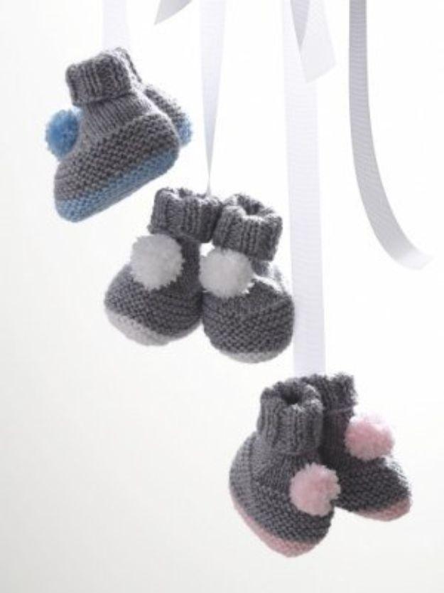 Three pairs of gray knitted baby booties with high cuffs and blue, white and pink pompoms, hanging on white ribbons