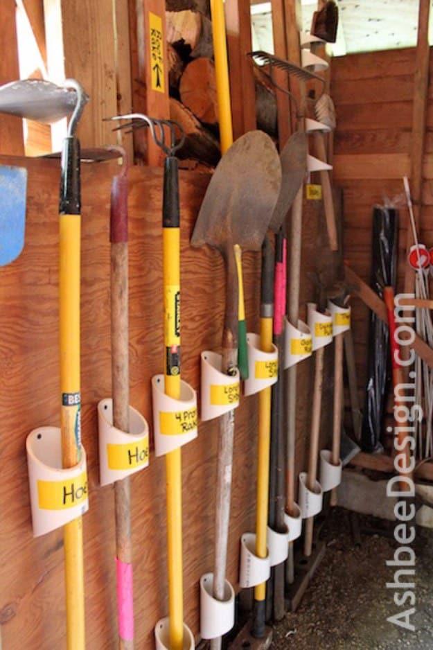 Long handled shovels and rakes standing in labeled white PVC pipe sleeves mounted to a rough wood shed wall