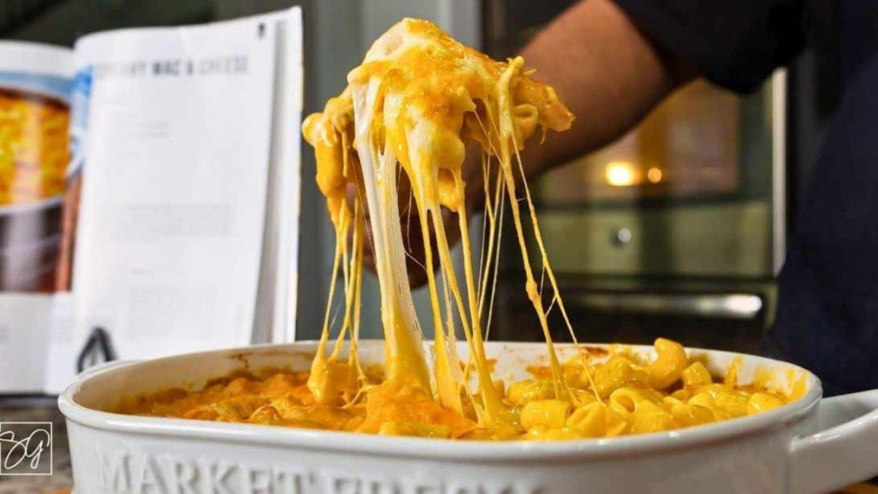 A hand lifting a long cheese pull from a white oval dish of elbow macaroni, a recipe page propped behind