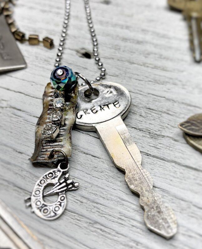 Vintage silver key stamped with the word create hanging on a ball chain with small charms and beads, on rustic wood