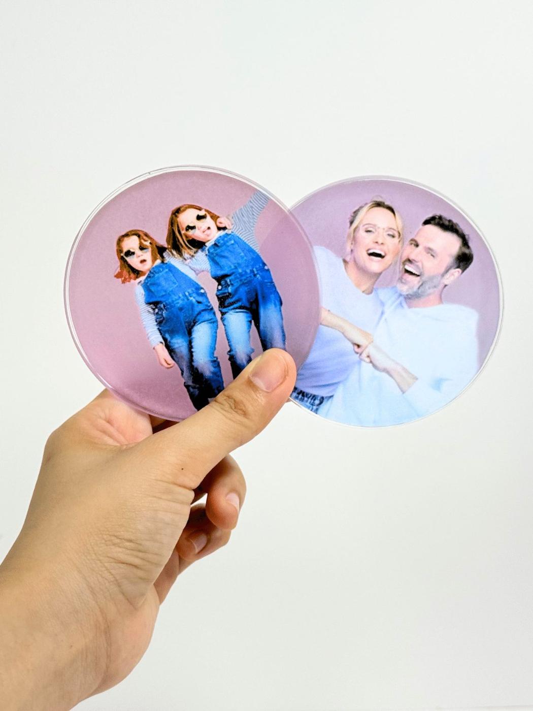 Hand holding two round resin coasters with a family photo and two kids in denim overalls sealed inside on a pink background