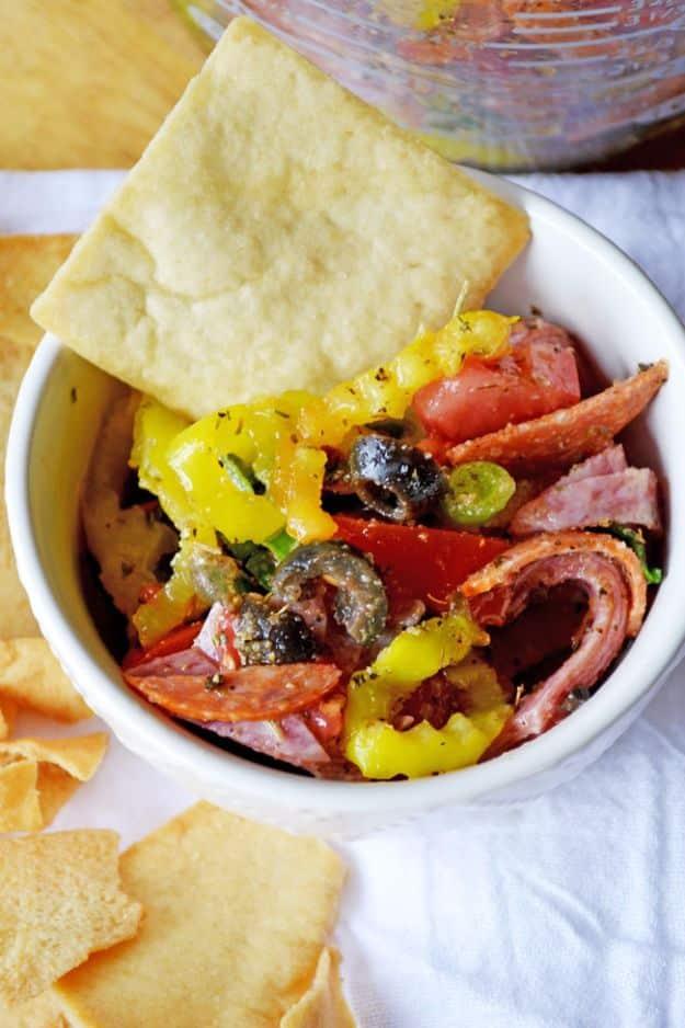 Chopped Italian dip in a white bowl with salami, pepperoni, banana peppers and black olives, pita chips around it
