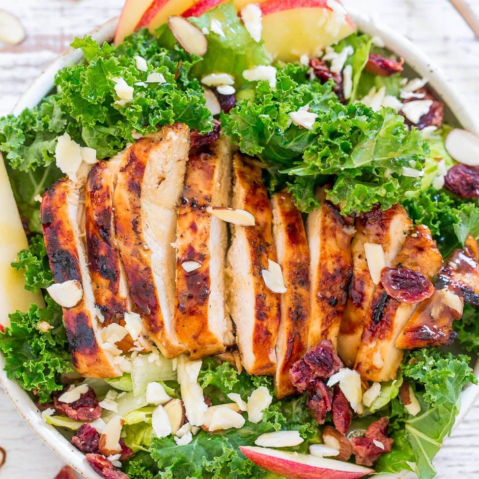Apple cheddar grilled chicken salad in a white bowl with kale, sliced grilled chicken, almonds and cranberries