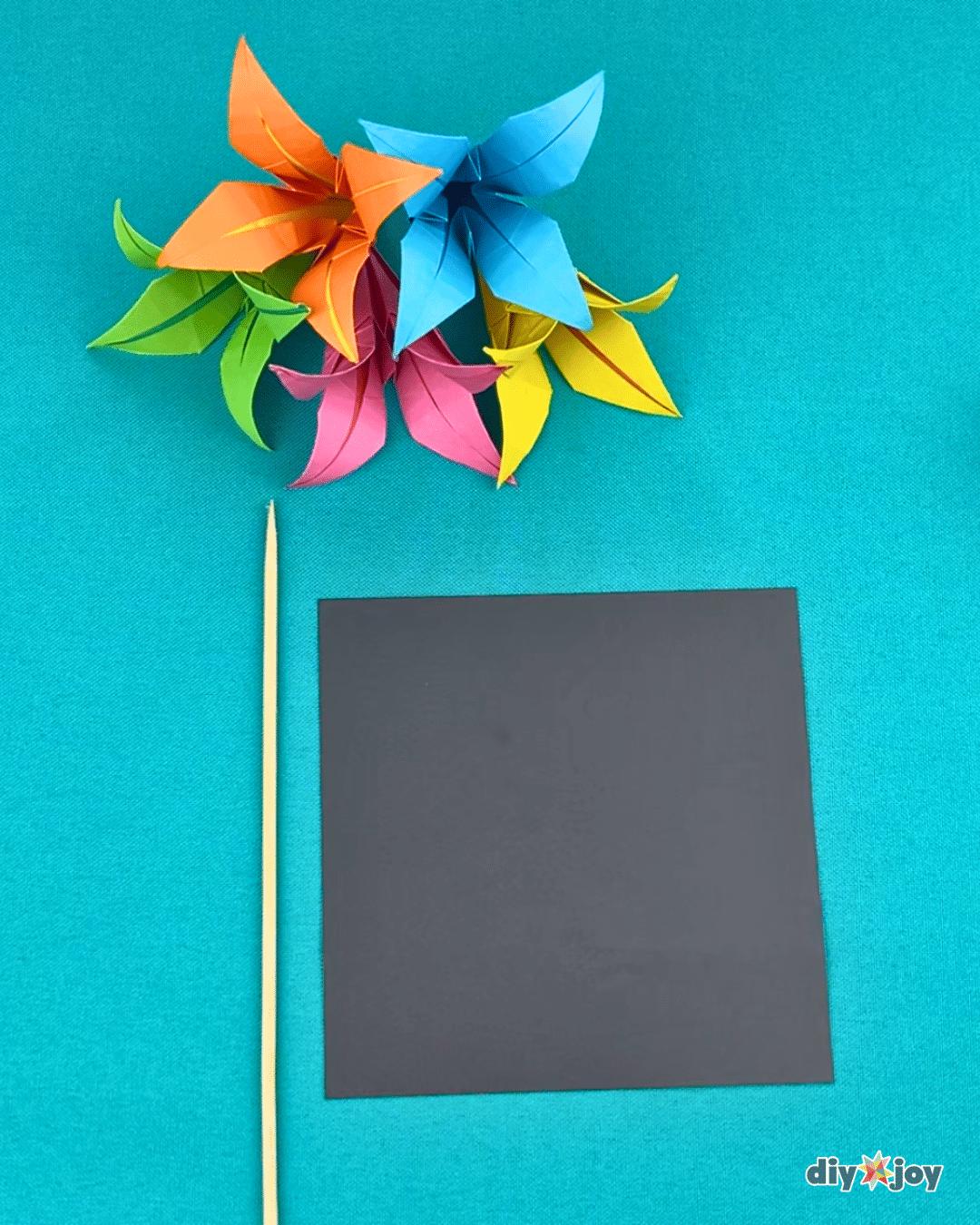 Paper lily materials laid out for the origami flower tutorial, square origami paper in several colors and a skewer