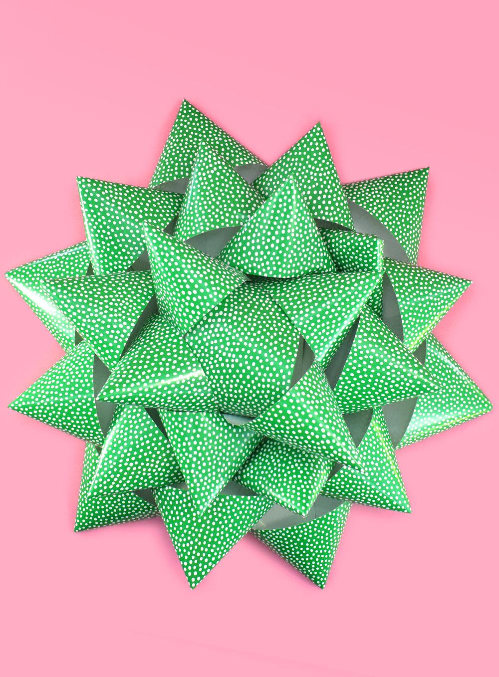 A huge handmade paper gift bow in green paper with white dots, about two feet across, on a pink background