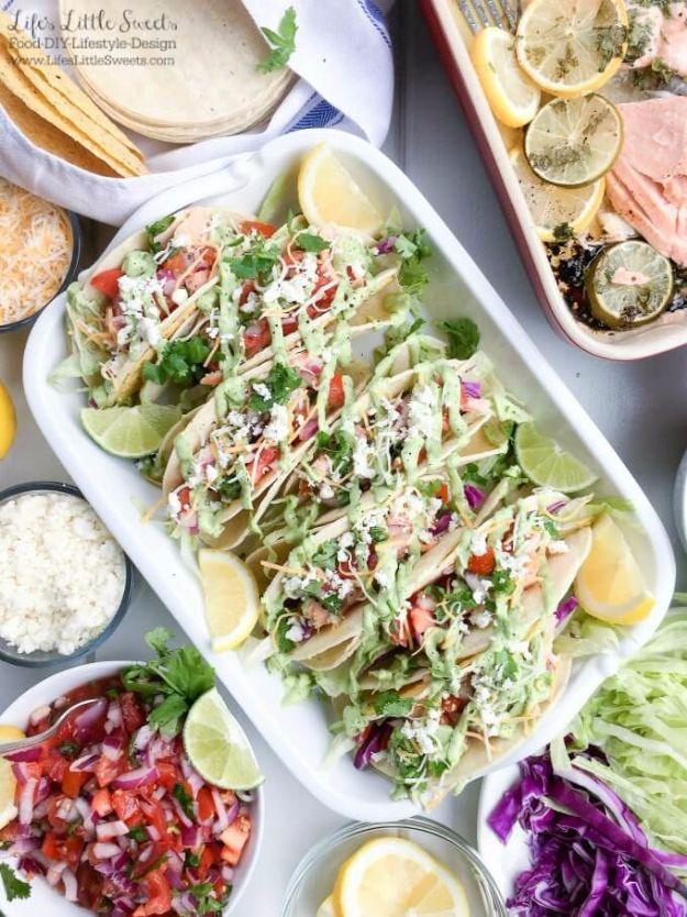 Salmon tacos on a white platter drizzled with avocado sauce, with bowls of fresh salsa, lime and cabbage