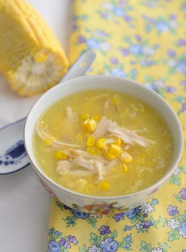 Chicken and sweetcorn soup in a floral bowl with shredded chicken and corn kernels, corn cob beside it
