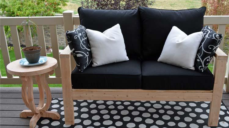 DIY outdoor loveseat in bare wood with black cushions and white pillows on a deck beside a small round side table