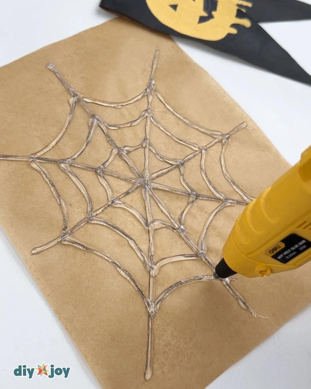 DIY Halloween Decor - a glue gun tracing the drawn web pattern in clear hot glue, with a paper bat nearby