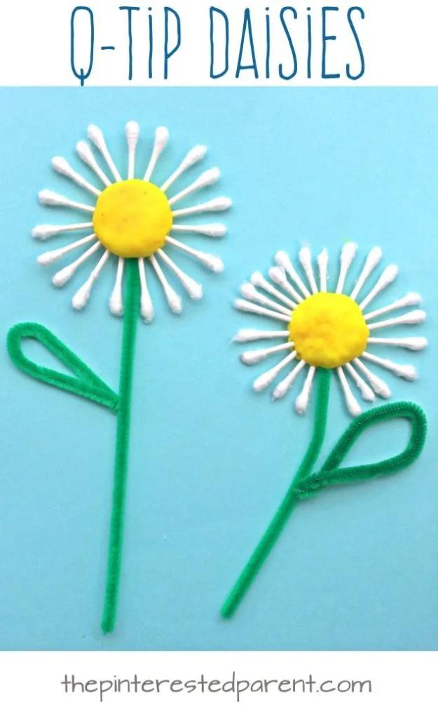 Two daisies of cotton swab petals around yellow play dough centers with green pipe cleaner stems, on blue