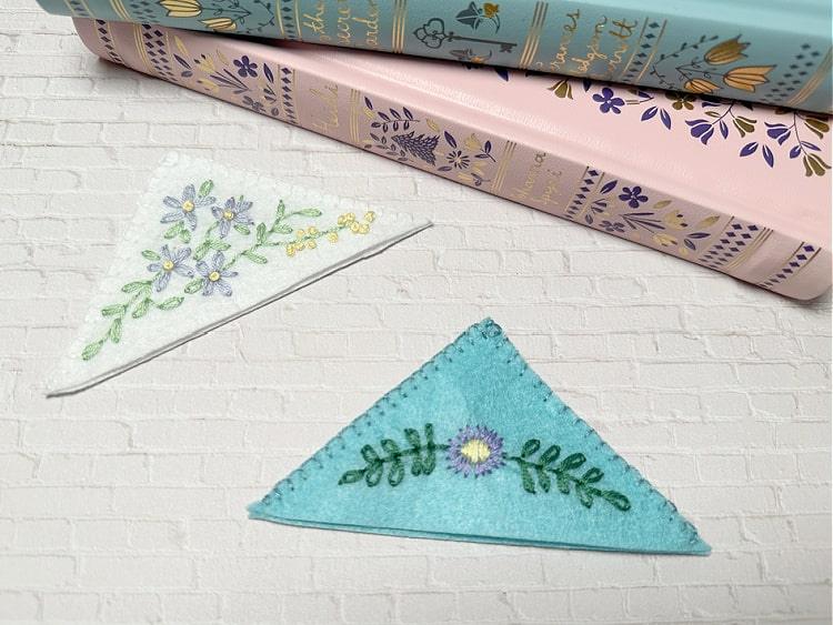 Two embroidered felt corner bookmarks, one cream with blue flowers and one teal with green leaves, on a notebook