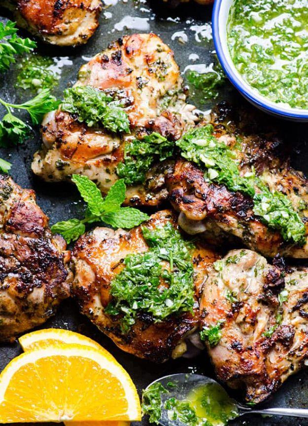 Grilled chimichurri chicken thighs topped with bright green parsley sauce on a dark pan with an orange slice