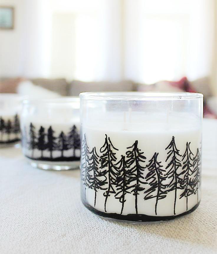 White jar candles hand drawn with a black paint marker pine treeline on a linen tablecloth with a sofa blurred behind them