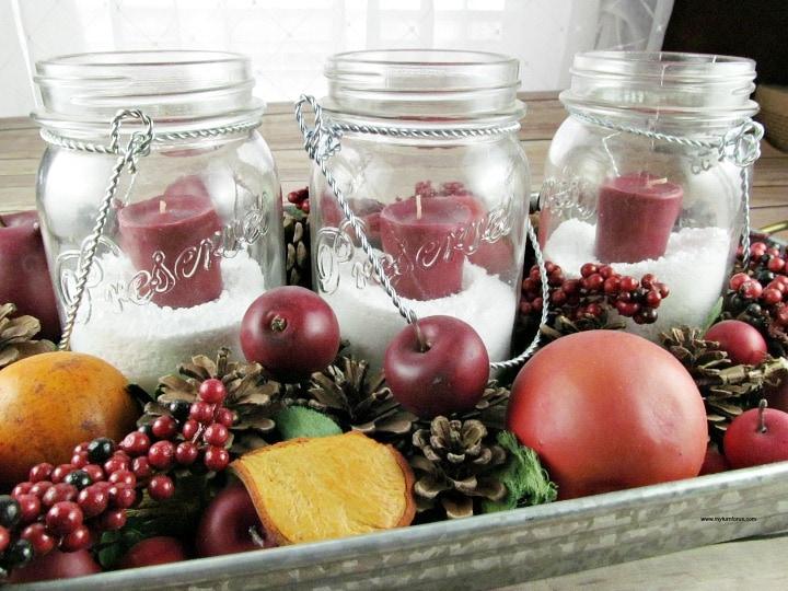 Three mason jars with Epsom salt snow and red votive candles on a metal tray surrounded by Christmas potpourri
