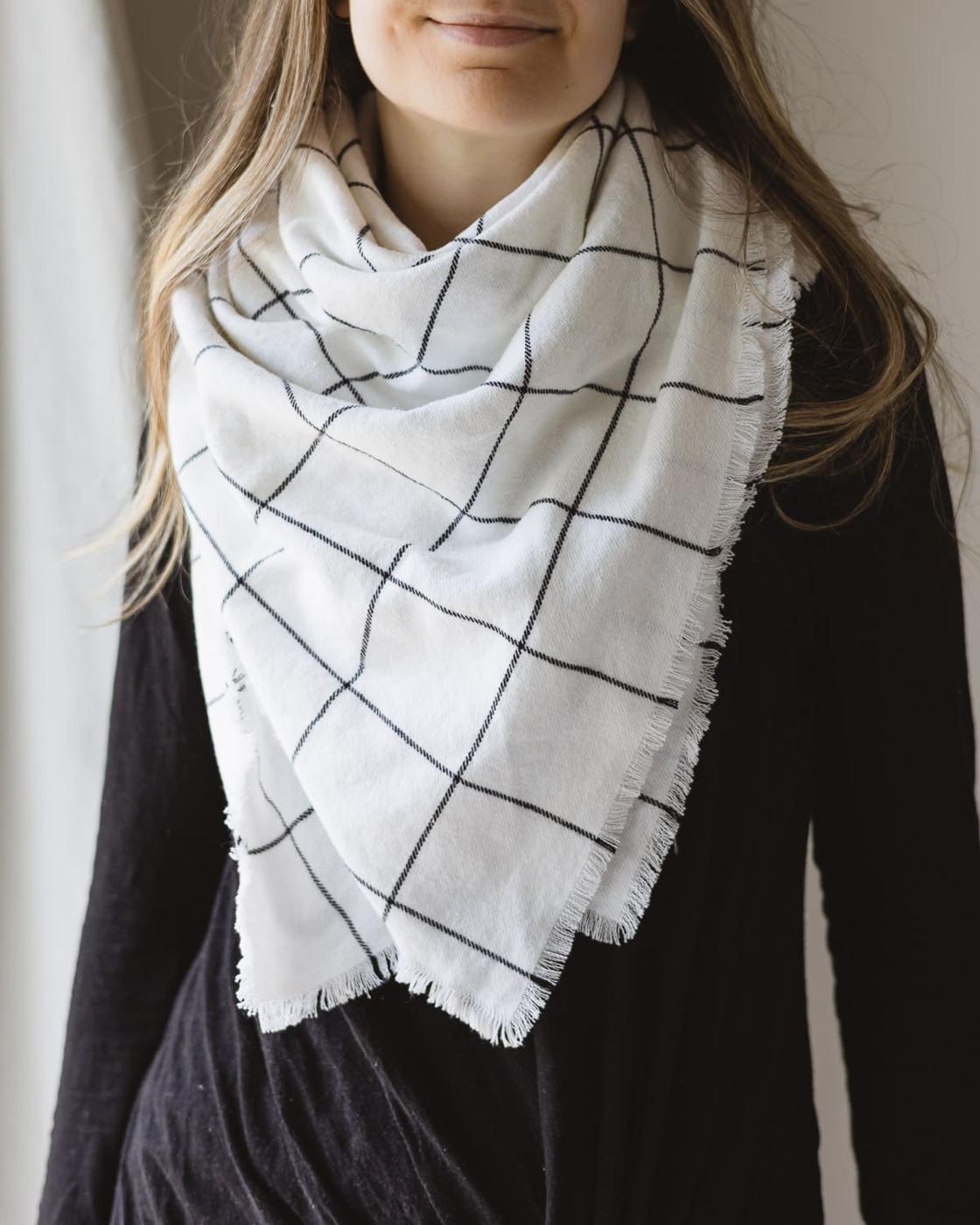 Woman in a black top wearing a fringed cream and black windowpane check flannel blanket scarf by a window