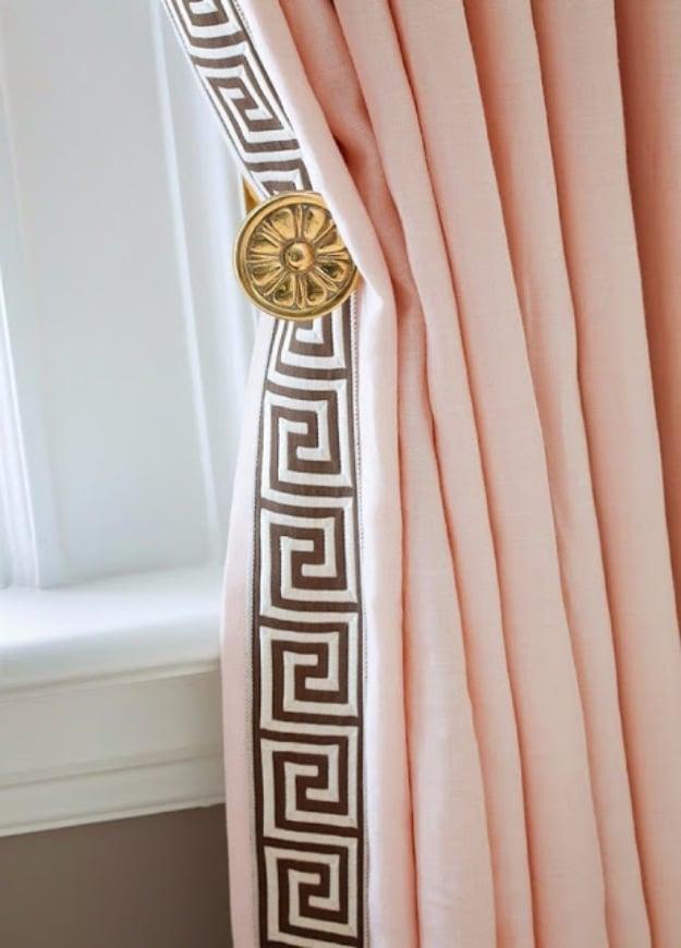 Blush pink linen curtain panel edged in brown and cream Greek key trim, held back by a gold rosette on a white wall