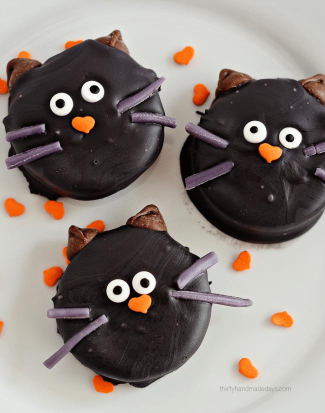 Three black Oreo cat cookies with candy eyes, pointed ears and whiskers arranged on a white plate for Halloween