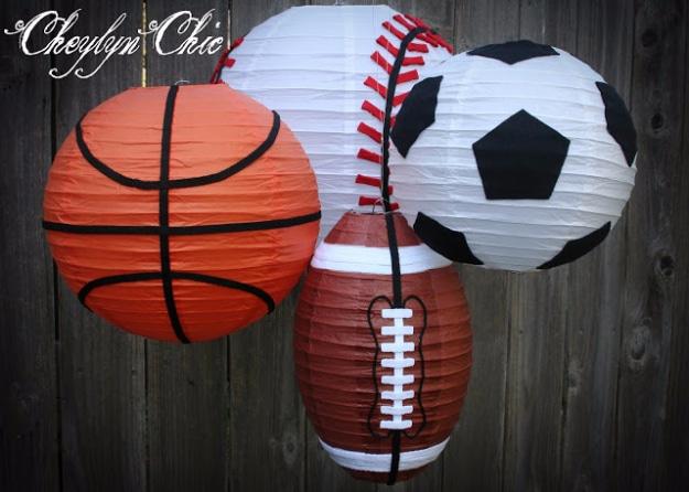 Four paper lanterns painted as a basketball, baseball, football and soccer ball, hung against a weathered fence