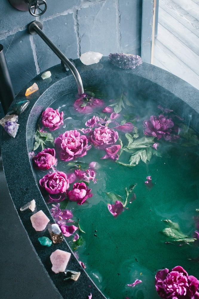 Spa like bath at home, a dark oval tub of green water with floating pink peonies and raw crystals set along the rim