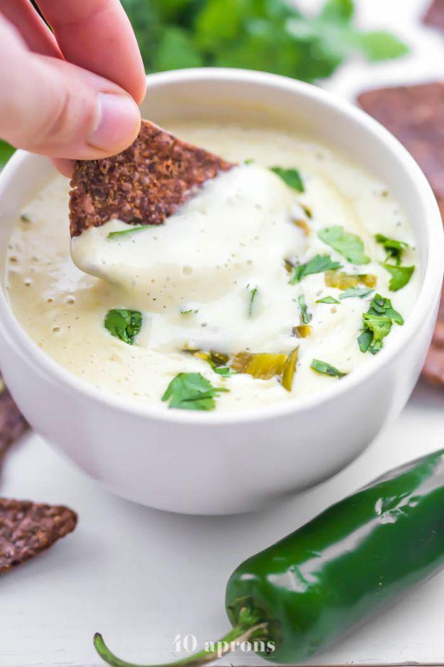 Hand dipping a dark grain tortilla chip into a white bowl of vegan queso blanco with cilantro and chilies