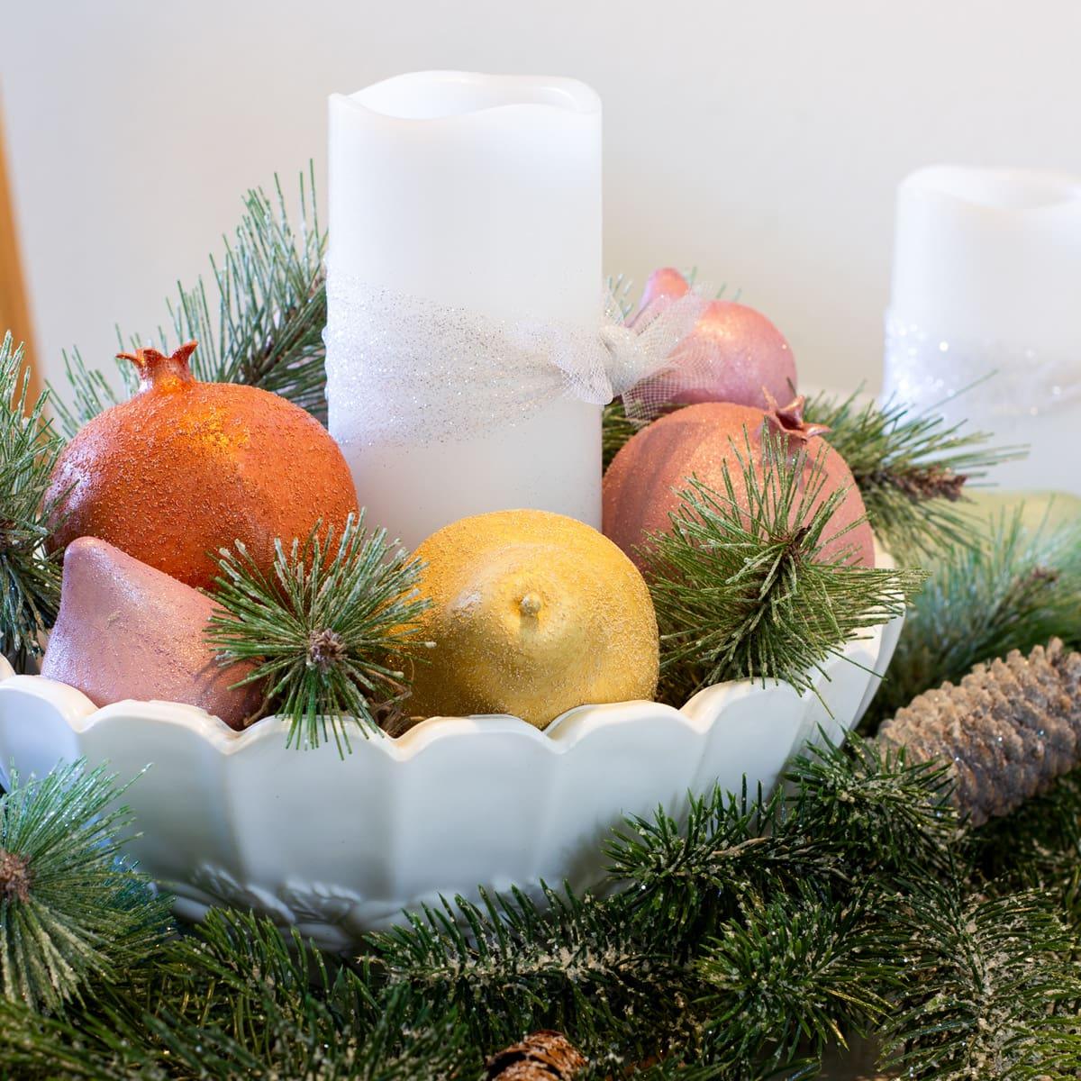 A white scalloped bowl with a pillar candle, glittery tulle and sugared faux pomegranates, pears and lemons in frosted pine