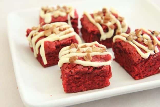 Five bright red velvet brownie squares on a white plate with pale drizzle and chopped golden pieces on top