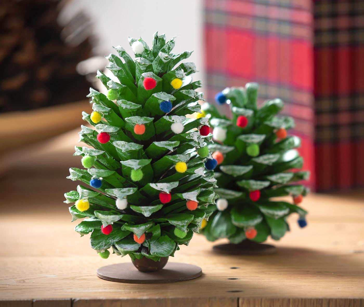 Pine cones painted green with white tipped scales and dotted with mini pom pom ornaments, standing on round wood bases