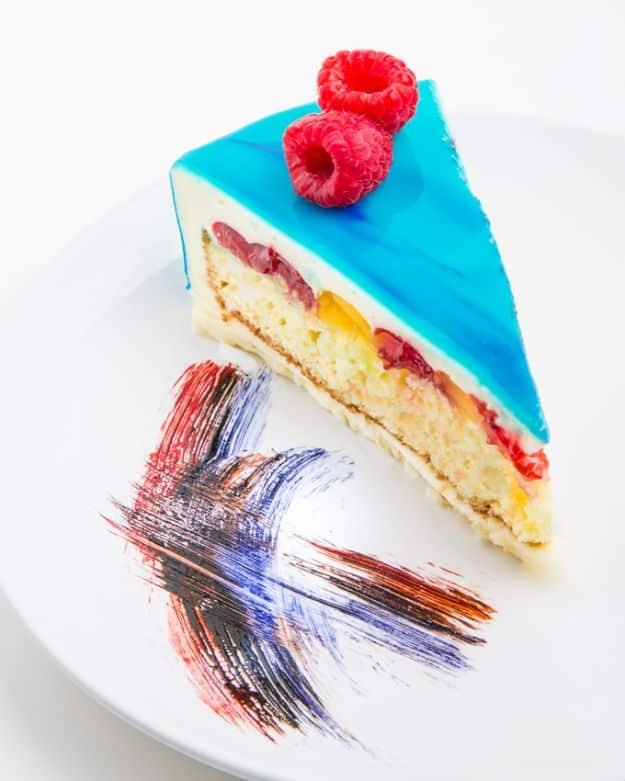 Slice of cake under a glossy bright blue mirror glaze topped with raspberries, with a painted streak on the plate