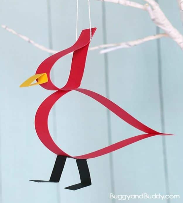 Red paper strip cardinal with black legs and a yellow beak, hanging on a string against a pale blue board wall