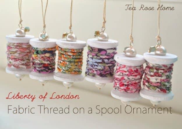 A wooden thread spool wrapped in floral fabric strips and cream lace, hung from twine against a pine branch