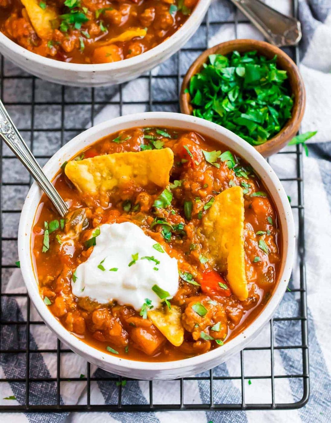Two white bowls of pumpkin chili with sweet potato, sour cream and tortilla chips standing in each, on a wire rack