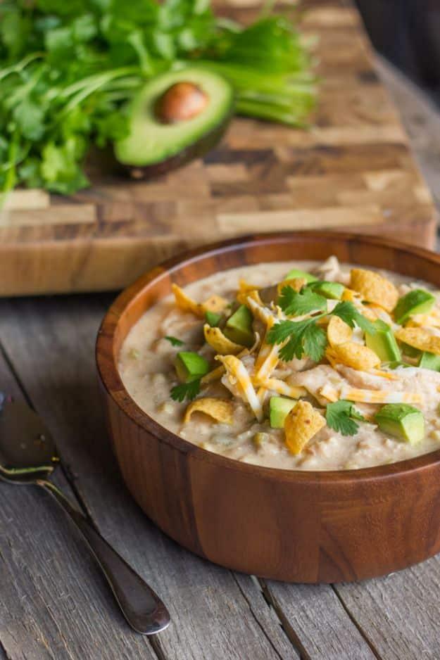 Creamy crockpot white chicken chili in a wooden bowl with diced avocado, cheese and scoop corn chips